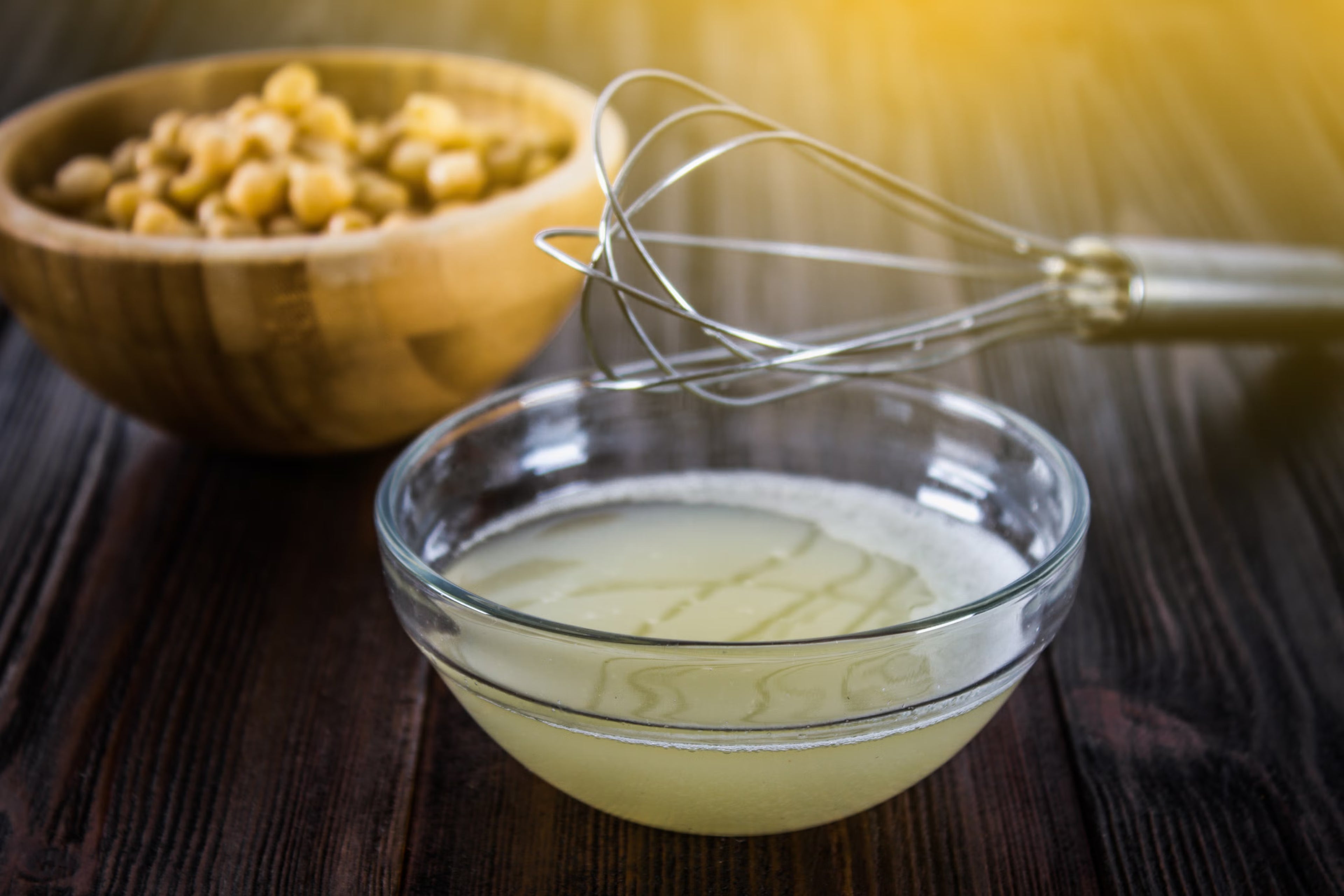 A small bowl containing liquid aquafaba with a whisk and a bowl of chickpeas in the background.
